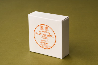 A white box labeled D.R. Harris Sandalwood Shaving Soap rests on a green surface. The label is circular, printed in orange with decorative text, and displays a weight of 100g. This luxurious shaving soap pairs classic sandalwood fragrance with sustainable, zero-waste grooming for an effortless shave.