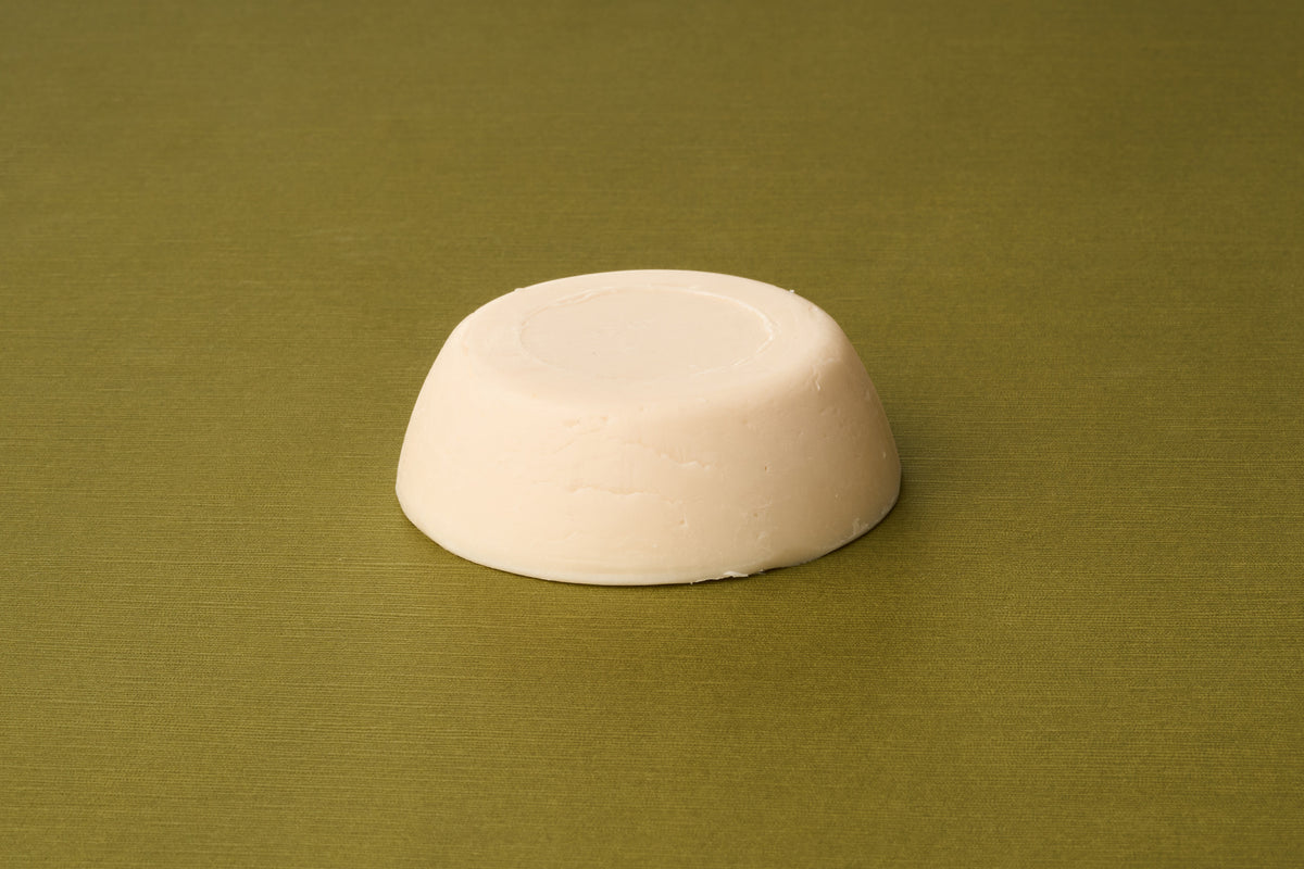 A round, solid, off-white bar of D.R. Harris Sandalwood Shaving Soap rests on a textured olive green surface. Featuring smooth sides and a flat top with a circular indentation at its center, this soap is an excellent option for those pursuing zero-waste shaving.