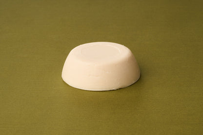 A round, solid, off-white bar of D.R. Harris Sandalwood Shaving Soap rests on a textured olive green surface. Featuring smooth sides and a flat top with a circular indentation at its center, this soap is an excellent option for those pursuing zero-waste shaving.