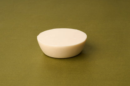The D.R. Harris Sandalwood Shaving Soap, a round, off-white bar, rests on a plain olive green surface and offers a zero-waste shaving experience.