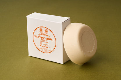 A round, off-white D.R. Harris Sandalwood Shaving Soap rests beside a white box labeled “Shaving Bowl Refill” on an olive green surface—an elegant choice for zero-waste shaving routines.