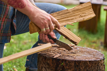 Using the Kershaw Camp 5, a person splits wood on a tree stump outdoors; wood chips and bark are scattered around, and the individual is wearing jeans and rings on their fingers.