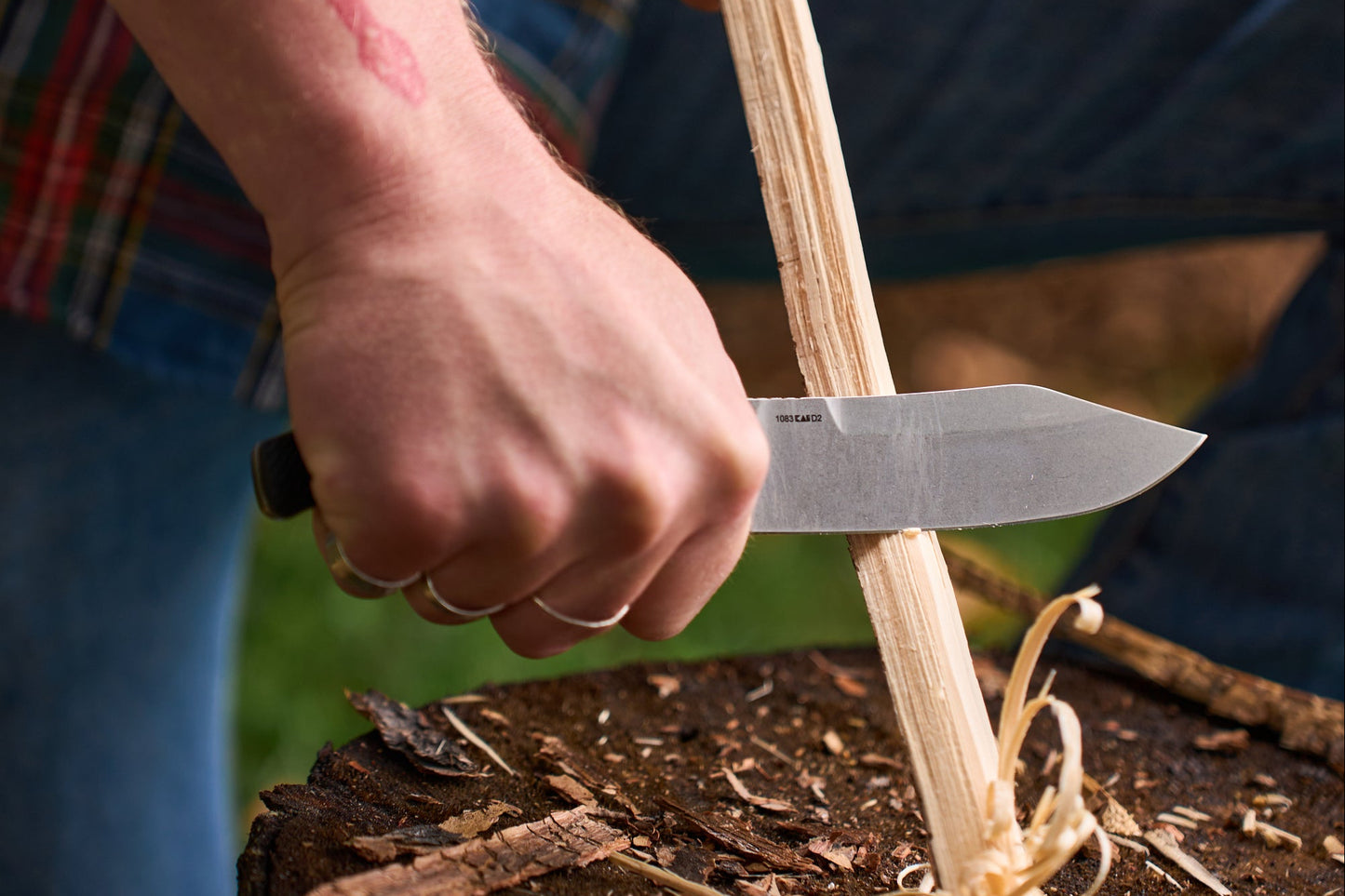 Person using a knife to cut wood strips on a wooden stump outdoors.