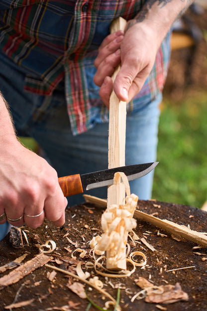A person in a plaid shirt uses the Helle Knives Viking 110mm Carbon Steel Hunting Knife to carve a piece of wood, creating wood shavings on a tree stump outdoors.