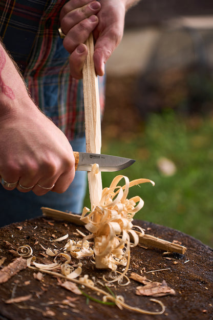Using the Helle Bleja, a person carves thin wood shavings from a stick, creating delicate curls. The close-up shows hands and the wood, set against a blurred green outdoor background.
