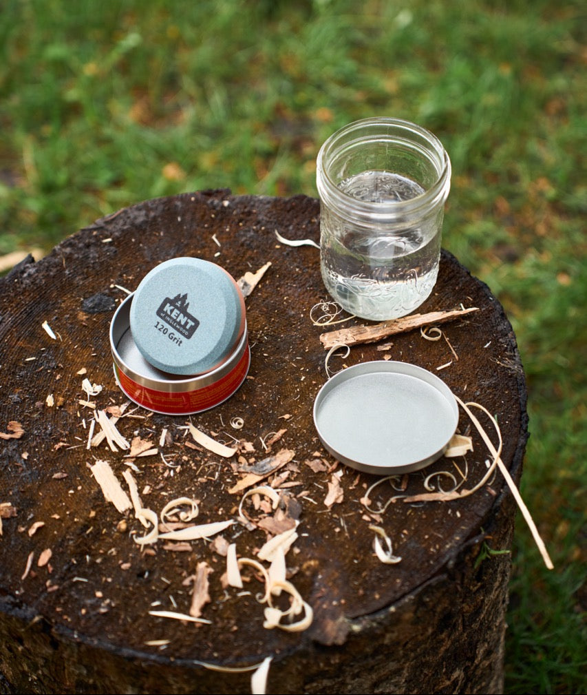 Camping utensils including a jar, metal cups, and wooden sticks on a log with grass in the background.