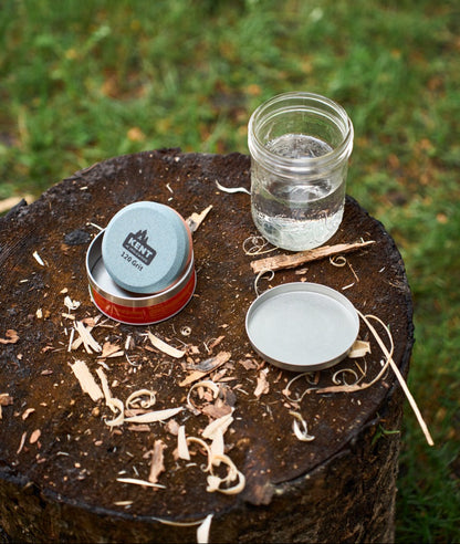 Camping utensils including a jar, metal cups, and wooden sticks on a log with grass in the background.
