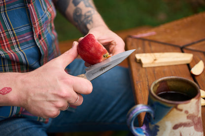 A person with tattoos peels a red apple at a wooden table using the RESQ Knives Korpi Camp Kitchen Knife, which is crafted in Finland from recycled stainless steel; a ceramic mug and apple slices sit nearby.