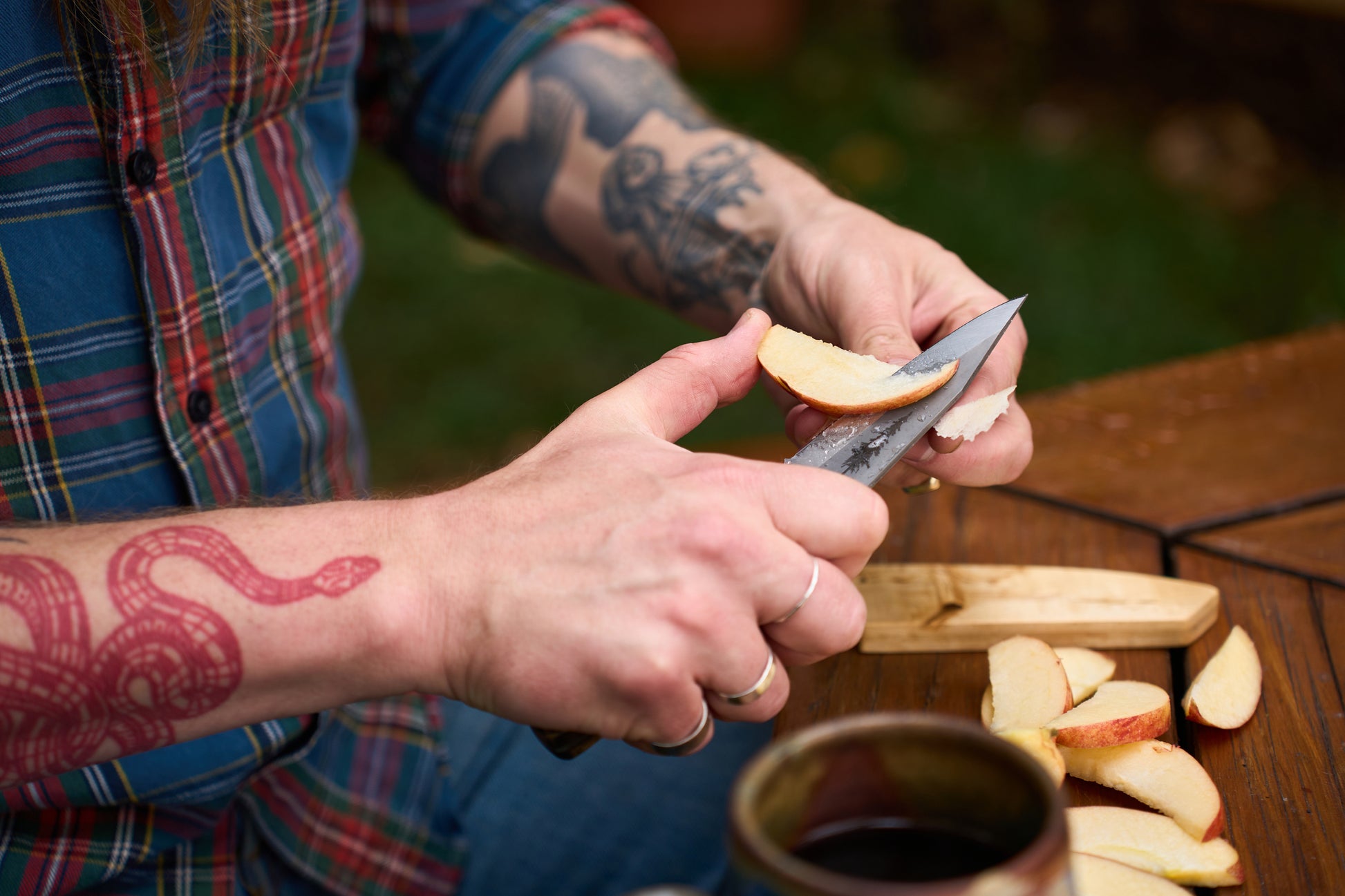 A person with tattoos slices an apple with the RESQ Knives Korpi Camp Kitchen Knife over a wooden board. Wearing a plaid shirt, they work at an outdoor wooden table where a mug rests nearby. The scene showcases the quality and durability of recycled stainless steel in the RESQ Knives Korpi, making outdoor food preparation both stylish and sustainable.