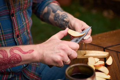 A person with tattoos slices an apple with the RESQ Knives Korpi Camp Kitchen Knife over a wooden board. Wearing a plaid shirt, they work at an outdoor wooden table where a mug rests nearby. The scene showcases the quality and durability of recycled stainless steel in the RESQ Knives Korpi, making outdoor food preparation both stylish and sustainable.