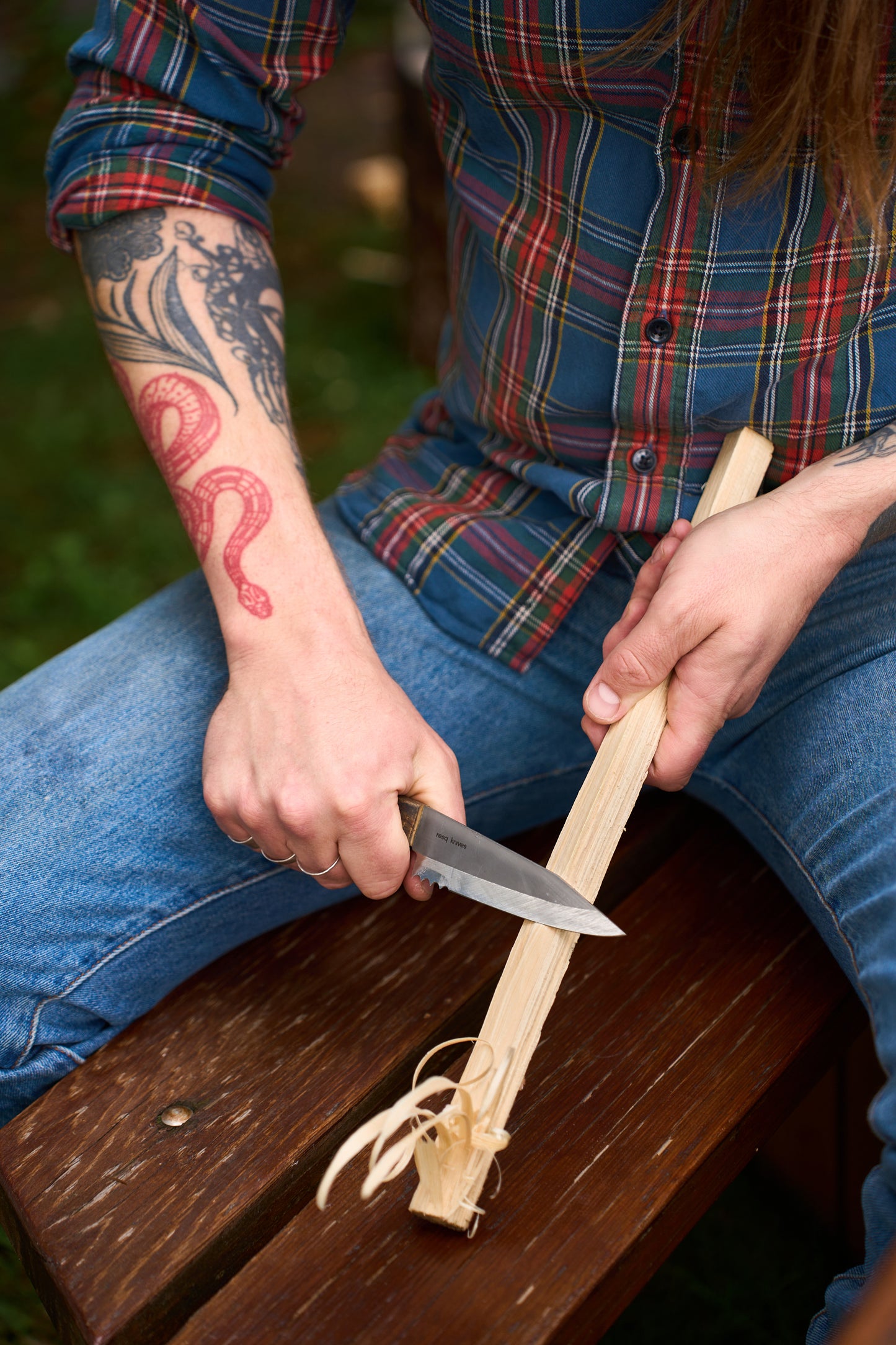 A person with a red snake tattoo is whittling a piece of wood with a RESQ Knives Korpi Camp Kitchen Knife made from recycled stainless steel, while sitting on a wooden bench, wearing blue jeans and a plaid shirt.