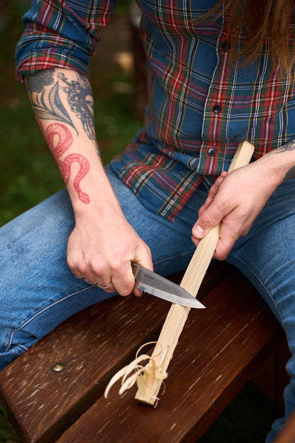 A person with a red snake tattoo is whittling a piece of wood with a RESQ Knives Korpi Camp Kitchen Knife made from recycled stainless steel, while sitting on a wooden bench, wearing blue jeans and a plaid shirt.
