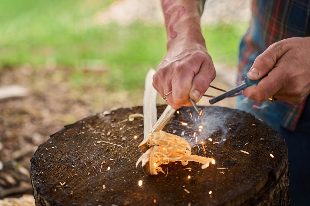 A person uses the Kupilka Firesteel on a tree stump to create sparks near wood shavings, with their tattooed arm and plaid shirt visible.