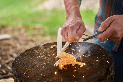 A person uses the Kupilka Firesteel on a tree stump to create sparks near wood shavings, with their tattooed arm and plaid shirt visible.