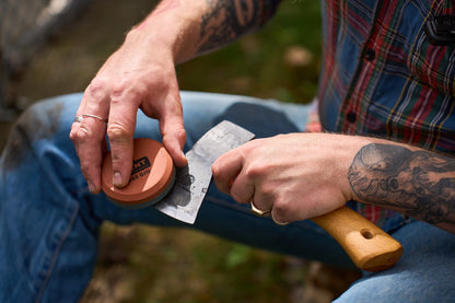 A tattooed person in jeans and a plaid shirt sharpens a small cleaver with a waterstone while sitting outdoors, possibly preparing for the Axe & Hatchet Sharpening Classes or getting ready to share expert tips on how to sharpen axes.