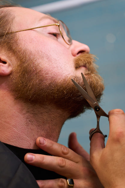 Wearing glasses, a person uses the Rockwell Hair and Beard Scissors to trim their beard, gently pulling the skin on their neck with one hand for precise and clean results with a neat, defined appearance.