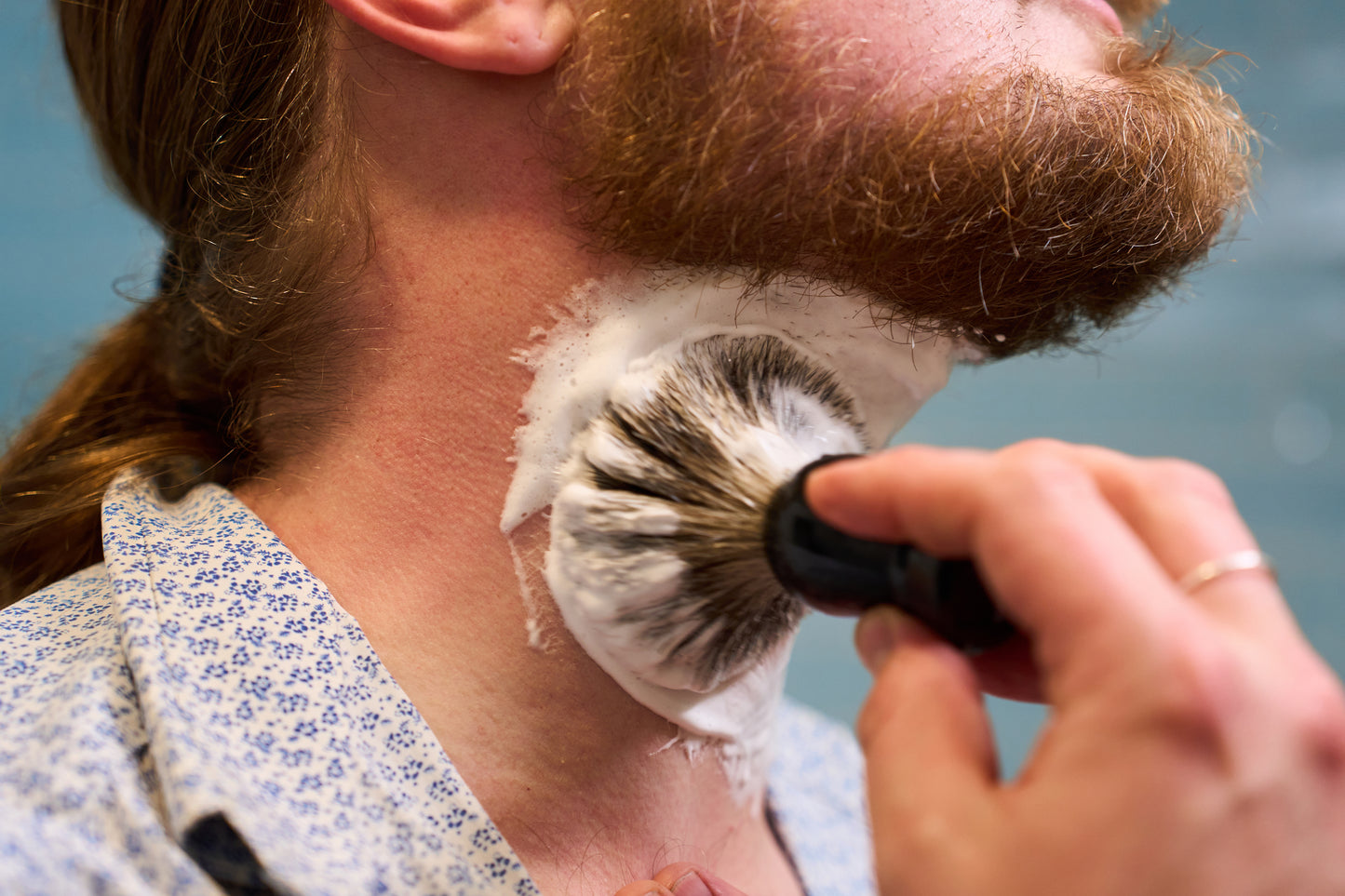Using the Classic Shaving Kit, a person with a red beard applies shaving cream to their neck with a badger shaving brush. Dressed in a blue and white patterned shirt, they prepare for a shave as part of their grooming routine.