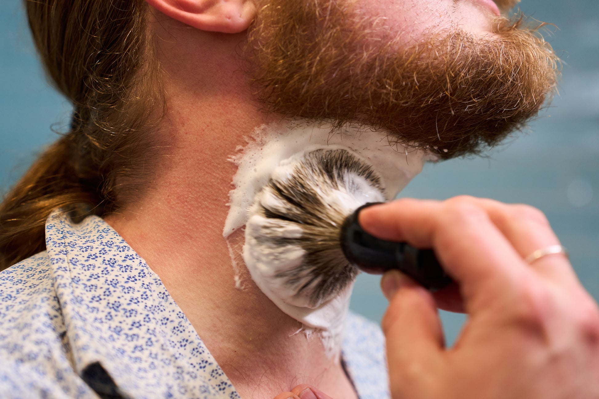 Using the Classic Shaving Kit, a person with a red beard applies shaving cream to their neck with a badger shaving brush. Dressed in a blue and white patterned shirt, they prepare for a shave as part of their grooming routine.