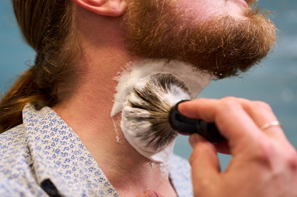 Using the Classic Shaving Kit, a person with a red beard applies shaving cream to their neck with a badger shaving brush. Dressed in a blue and white patterned shirt, they prepare for a shave as part of their grooming routine.