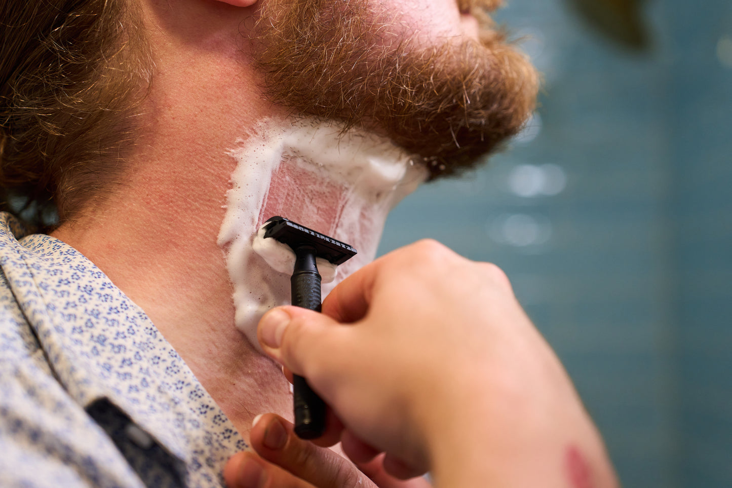 A close-up of a person using the Classic Shaving Kit’s black safety razor to shave their neck, removing shaving cream and hair below their beard while wearing a patterned shirt.