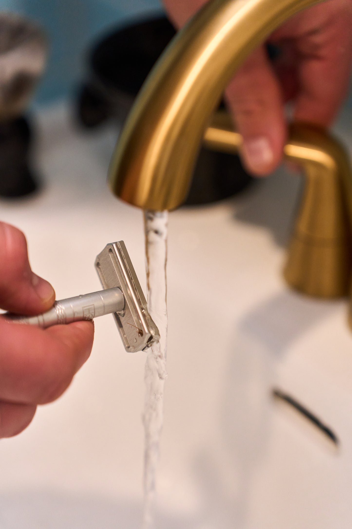 A close-up of hands rinsing the Henson Safety Razor Aircraft Aluminum under a stream of water from a gold-colored bathroom faucet above a white sink.