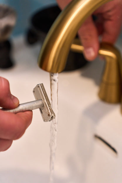 A close-up of hands rinsing the Henson Safety Razor Aircraft Aluminum under a stream of water from a gold-colored bathroom faucet above a white sink.