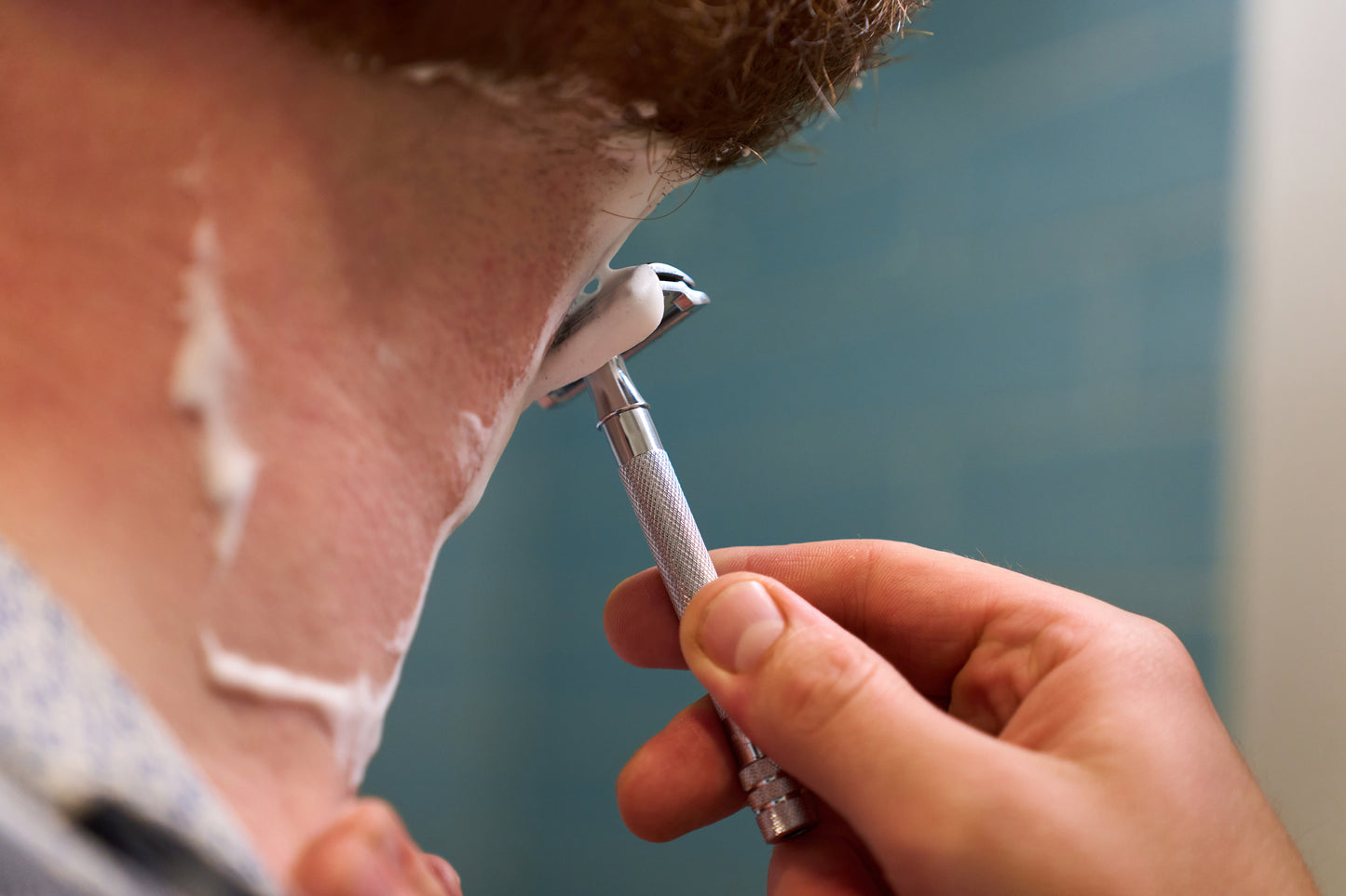 A close-up of a person shaving the back of their neck with the Merkur Chrome DE Safety Razor, cream applied, in a bathroom with blue tiles.