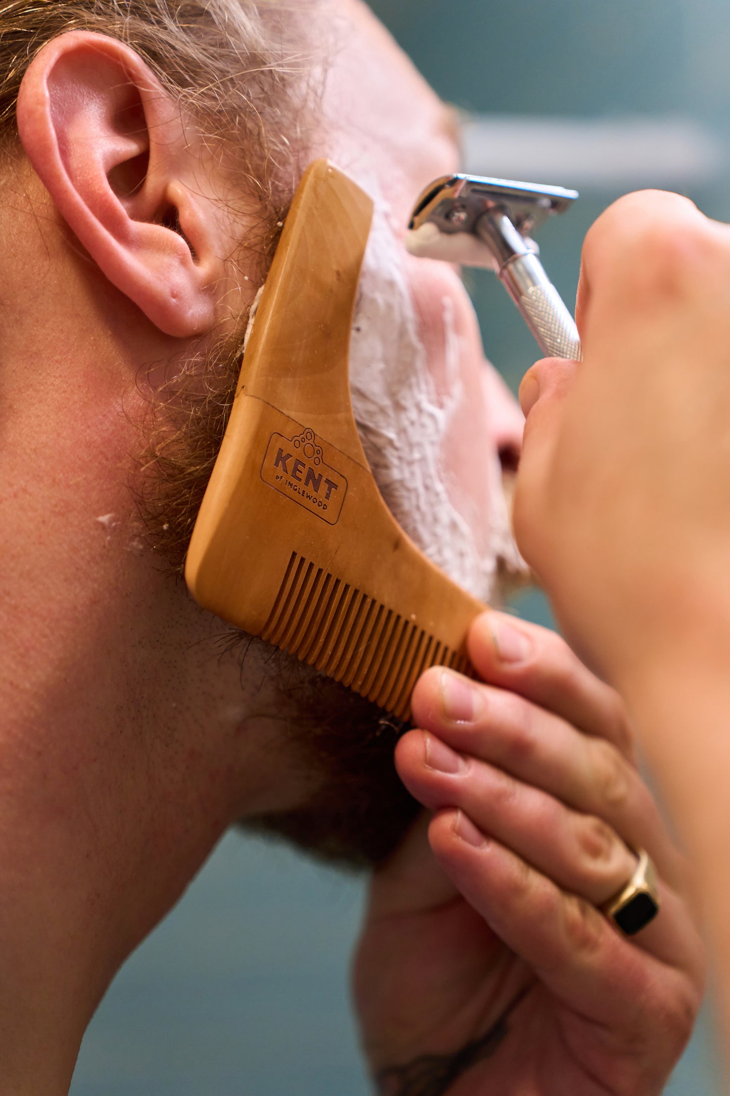 A person shaves their face with a razor, using the Kent of Inglewood Beard Shaper to guide the shave. Their cheek is covered in shaving cream as they hold the beard shaper to their jawline for precision.