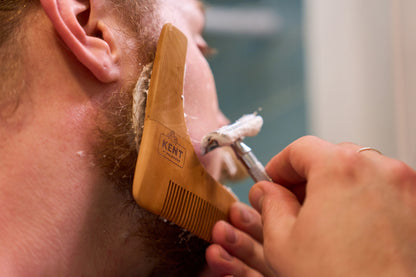 A person uses the Kent of Inglewood Beard Shaper and a razor to shave and define the edge of their beard, applying shaving cream along the jawline. The Kent of Inglewood Beard Shaper serves as a precise beard trimming tool, helping to shape and maintain clean lines for a well-groomed look.