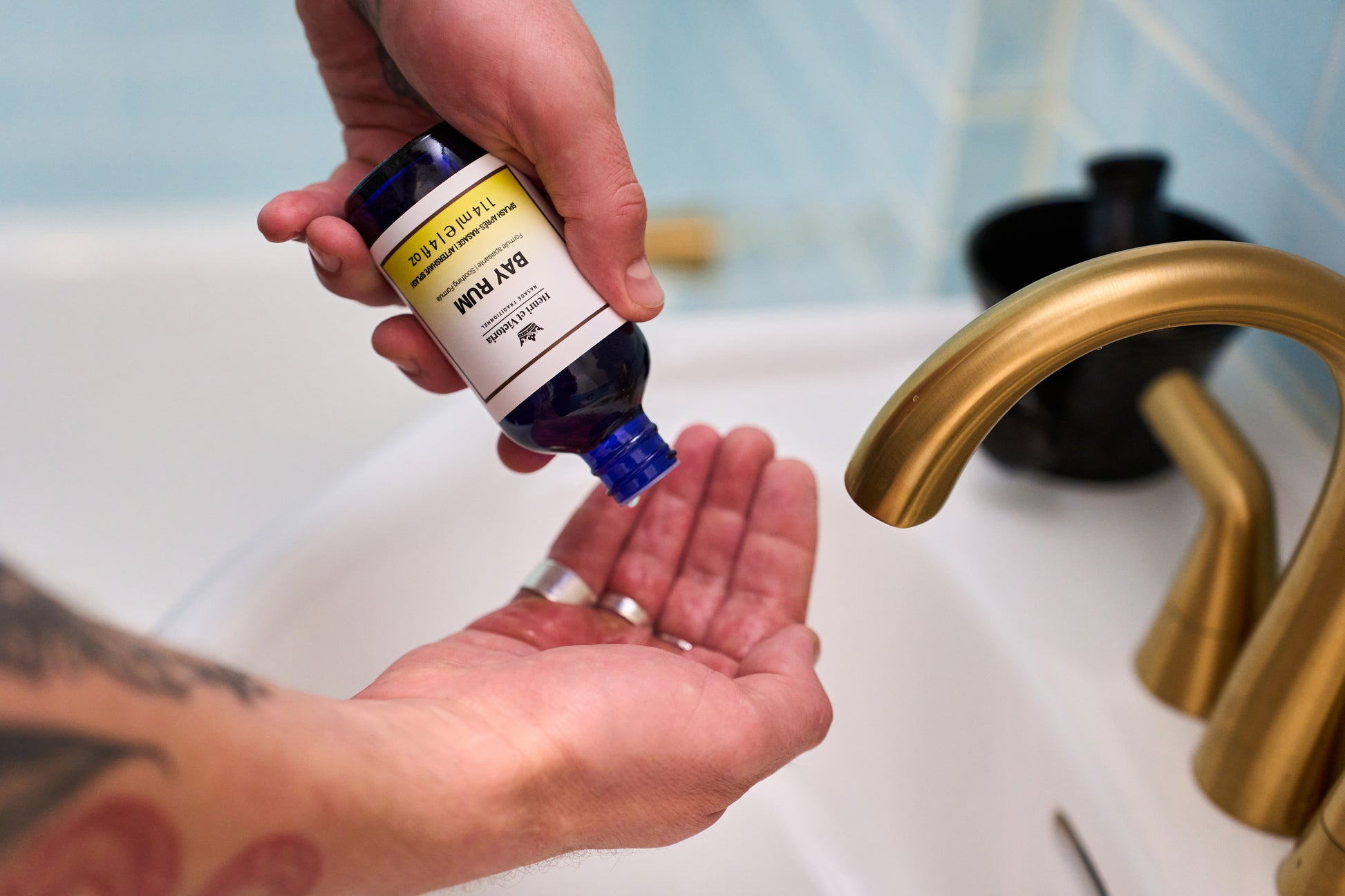 A person pours liquid from a blue bottle labeled "Henri et Victoria - Bay Rum Aftershave Splash" into their hand over a white sink with a gold faucet, preparing to enjoy an invigorating aftershave experience.