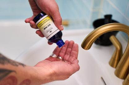 A person pours liquid from a blue bottle labeled "Henri et Victoria - Bay Rum Aftershave Splash" into their hand over a white sink with a gold faucet, preparing to enjoy an invigorating aftershave experience.