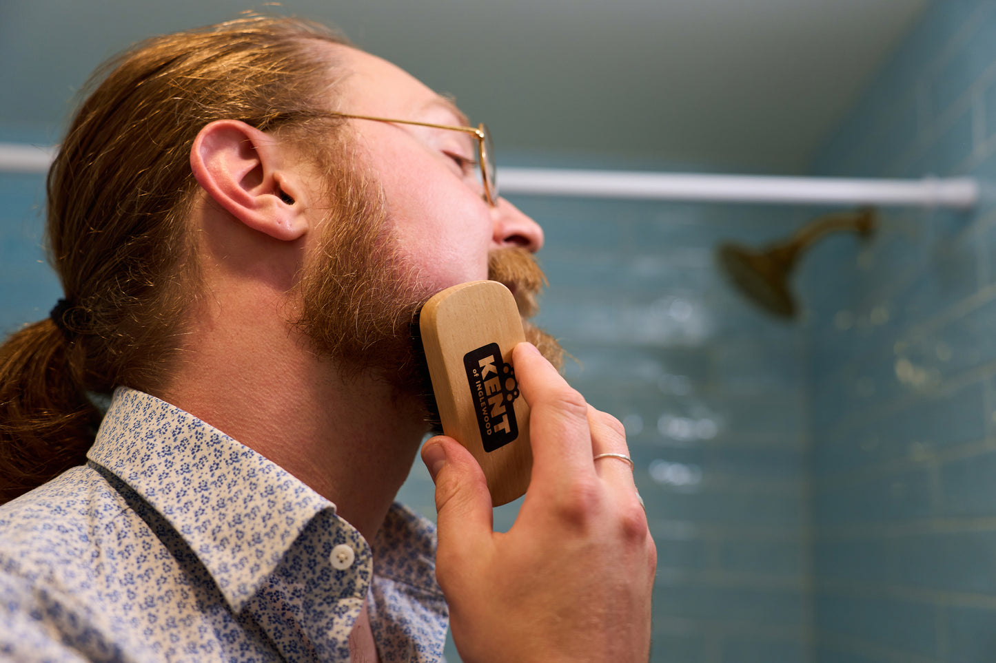A person with glasses and a patterned shirt grooms their beard with a Kent Of Inglewood Boar Bristle Beard Brush, practicing dedicated beard care in a bathroom featuring blue tiled walls and a showerhead in the background.