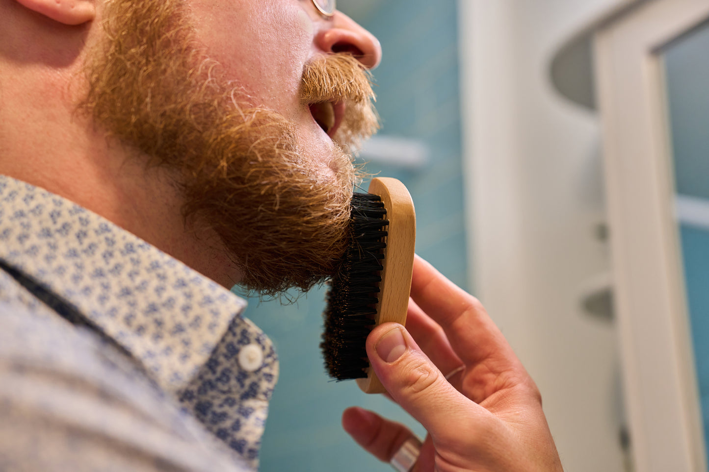A person with a reddish beard stands in front of a mirror, carefully grooming their beard with the Kent Of Inglewood Boar Bristle Beard Brush.