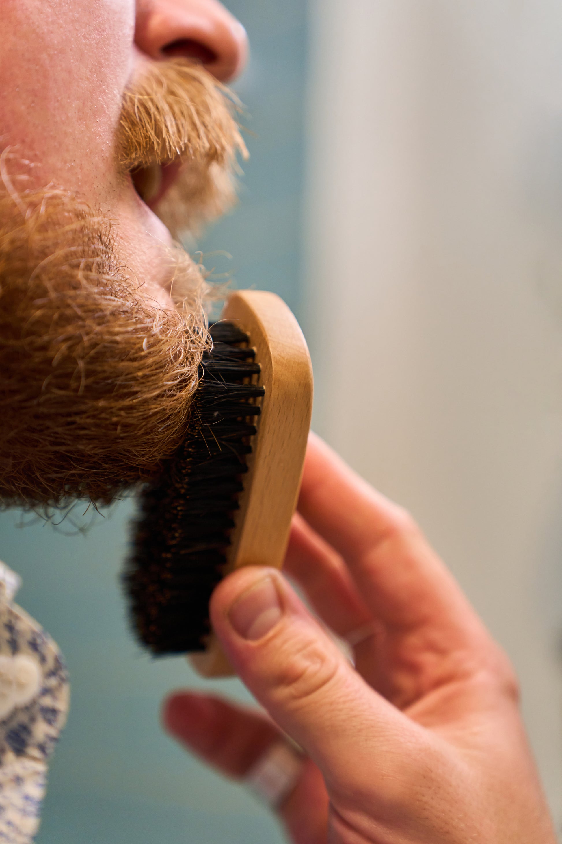 A close-up of a person brushing their reddish-brown beard with the Kent Of Inglewood Boar Bristle Beard Brush, its wooden handle in hand, highlighting part of their mouth and mustache as they focus on grooming their beard.