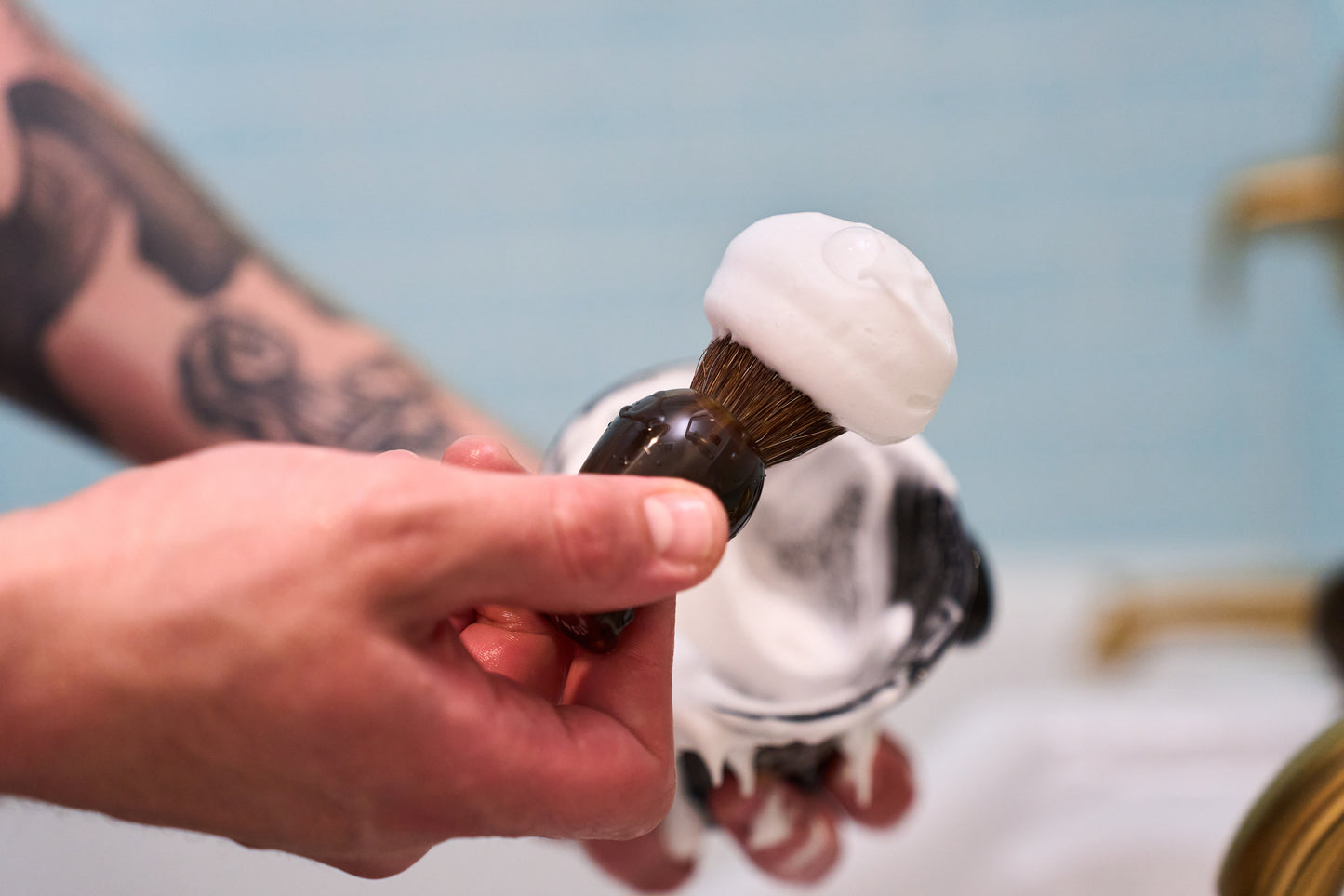 A person with a tattooed arm holds a Kent of Inglewood Horse Hair Shaving Brush topped with white shaving cream over a bowl, preparing for a shave.