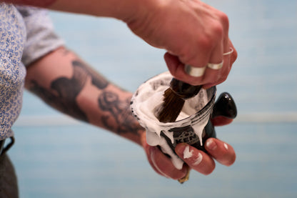 A person with tattoos uses the Kent of Inglewood Horse Hair Shaving Brush to mix shaving cream in a shaving bowl, holding the bowl in one hand. The softly blurred background contrasts with the focus on their hands.