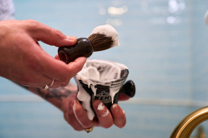 A person swirls the Kent of Inglewood Horse Hair Shaving Brush in a cup filled with shaving cream, with some foam on their fingers. The background features light blue tiles.