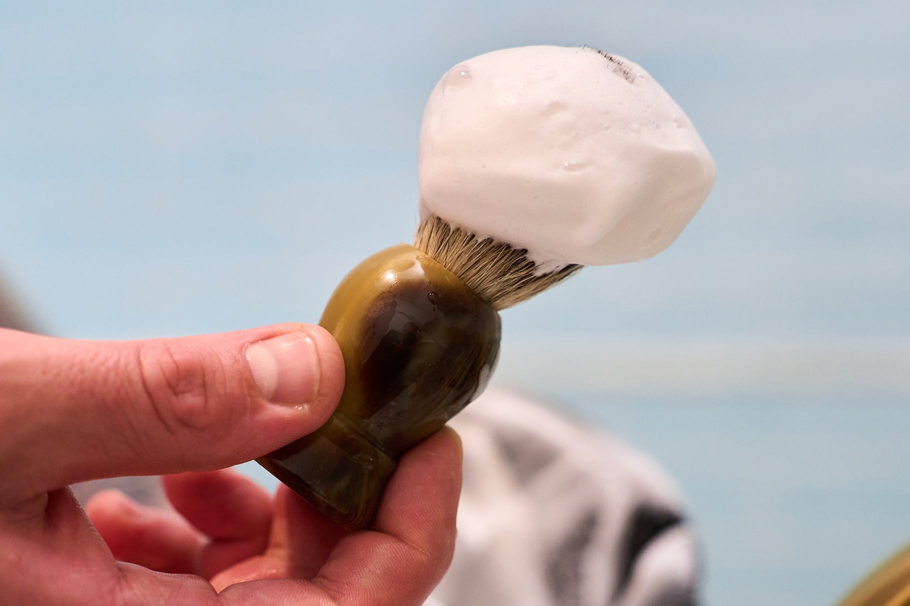 A hand holding a Kent of Inglewood Grey Badger Hair Shaving Brush with a brown handle and bristles covered in thick white shaving foam, perfect for lathering hard soaps, set against a blurred blue and white background.