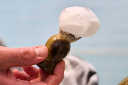 A hand holding a Kent of Inglewood Grey Badger Hair Shaving Brush with a brown handle and bristles covered in thick white shaving foam, perfect for lathering hard soaps, set against a blurred blue and white background.
