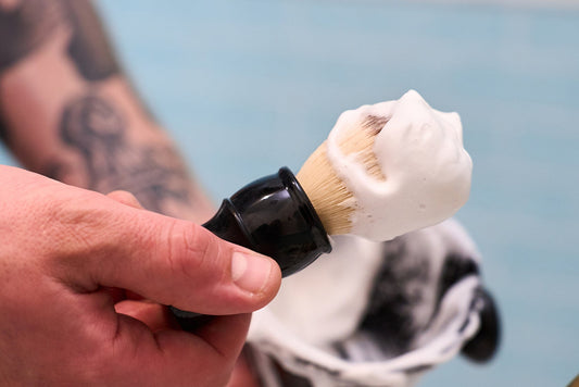 A hand holding the Kent of Inglewood Synthetic Fibre Shaving Brush covered in white shaving cream, with a bowl of shaving cream in the background.