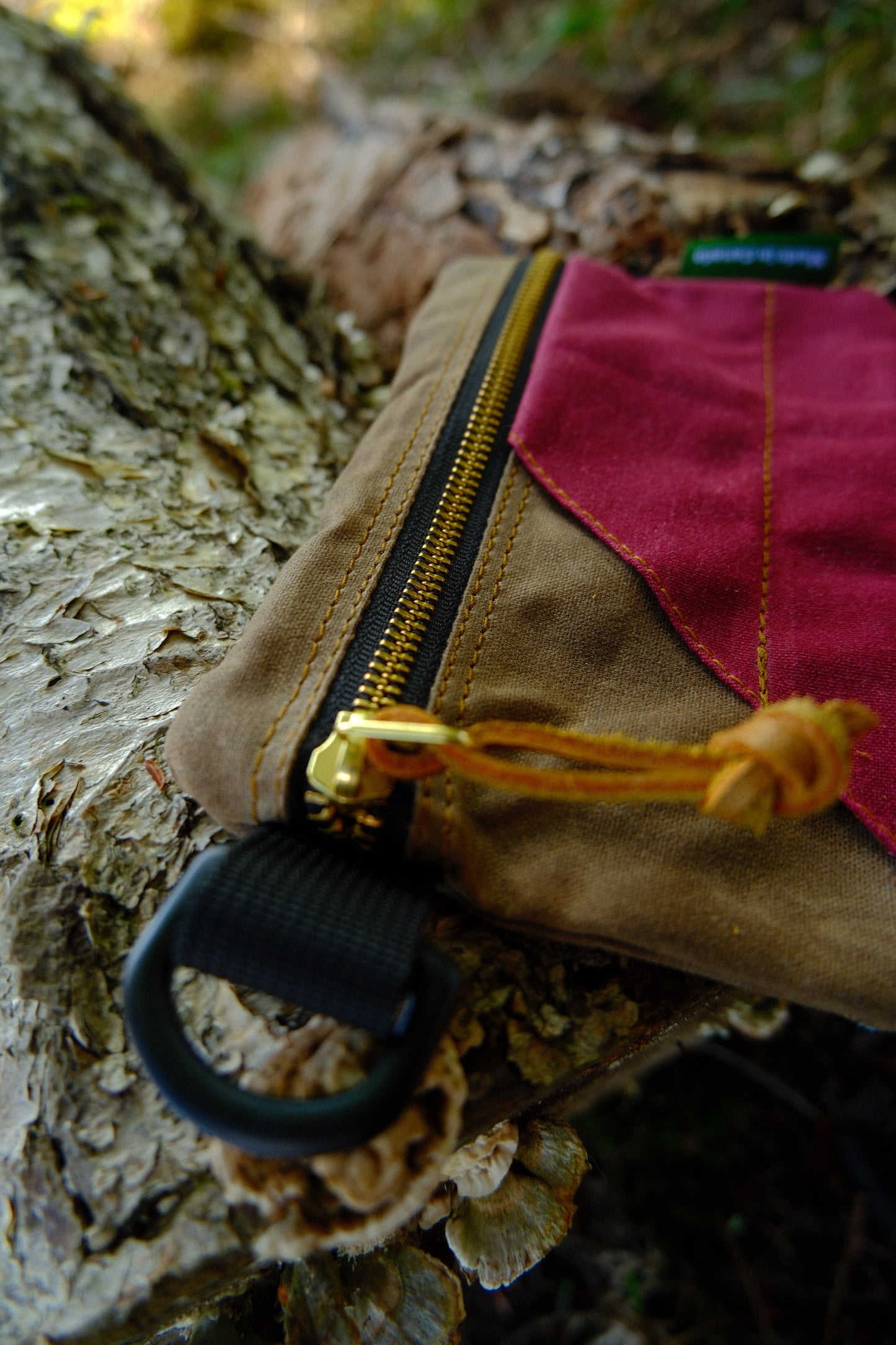 A close-up of the Bowman Gear Sentry Waxed Canvas EDC Pouch—featuring brown and red waxed canvas, a black zipper, and a yellow pull cord—designed for essentials storage, resting outdoors on a textured tree trunk.
