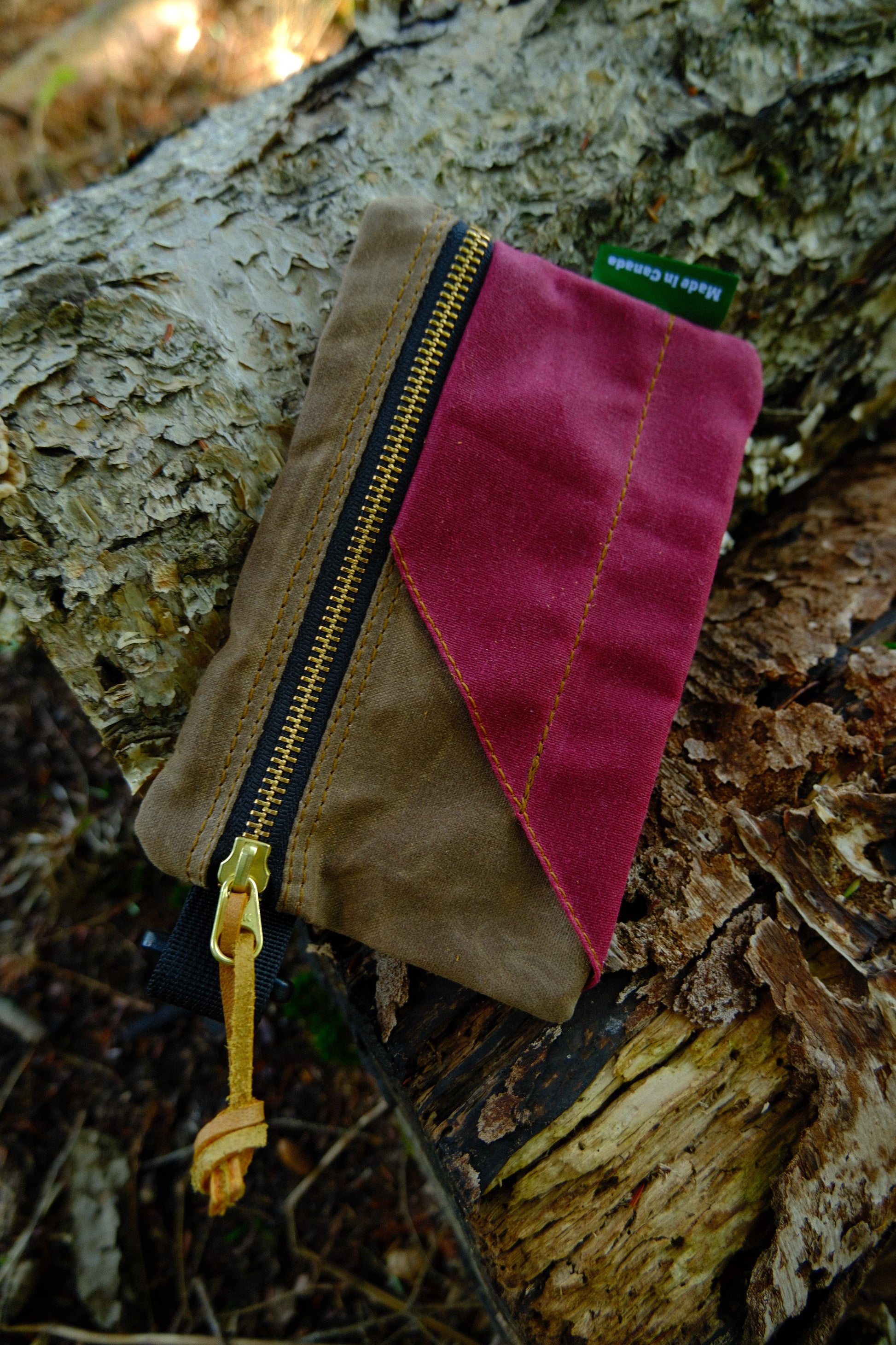 The Bowman Gear Sentry Waxed Canvas EDC Pouch—a compact pouch with a brown body, red accent, and gold zipper—rests on a rugged tree trunk outdoors. A green tag stating “Made in Canada” highlights its quality craftsmanship and versatile essentials storage.
