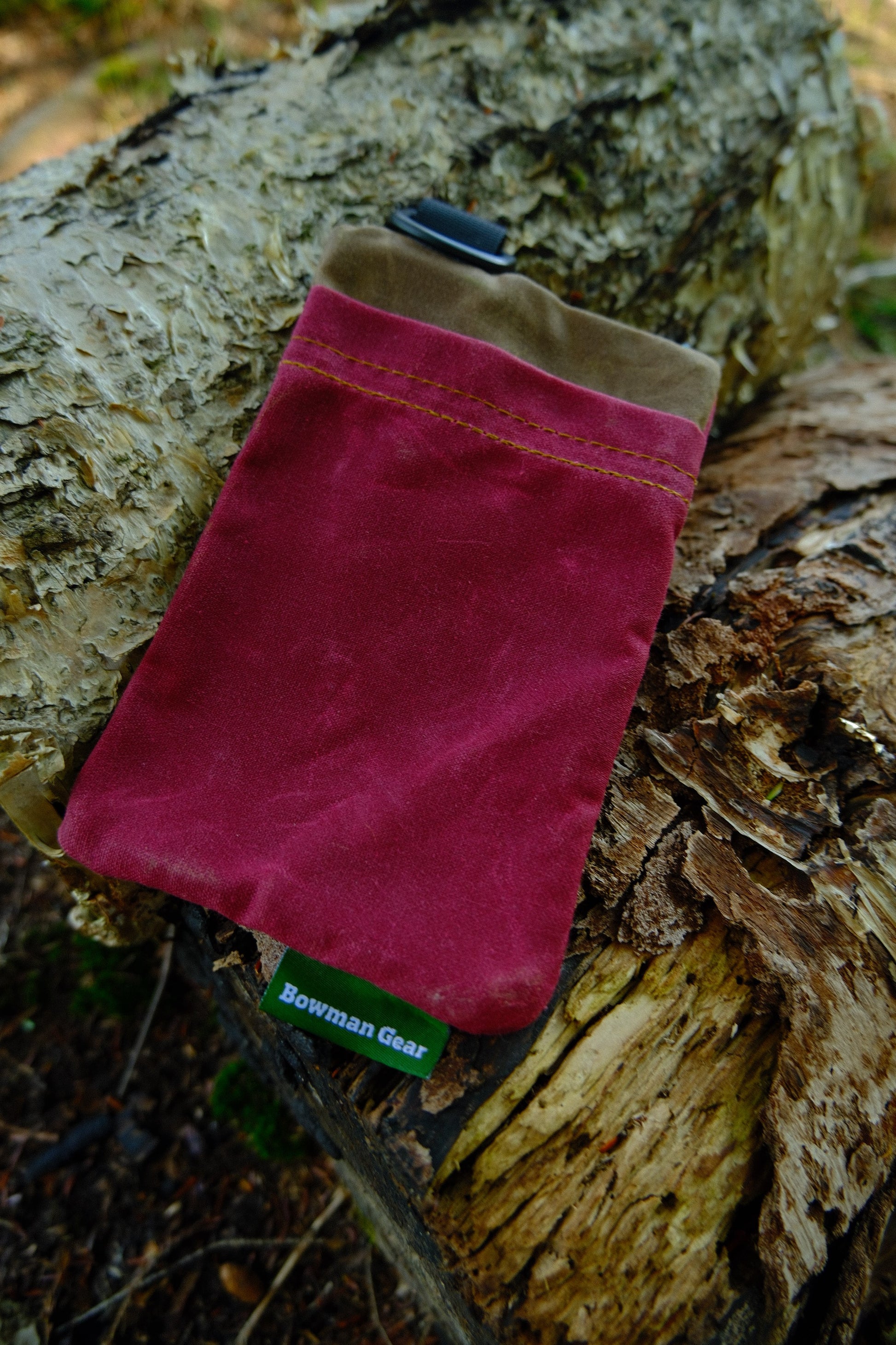 A red and brown Bowman Gear Sentry Waxed Canvas EDC Pouch sits atop a weathered tree log outdoors, providing rugged storage for essential items on any adventure.