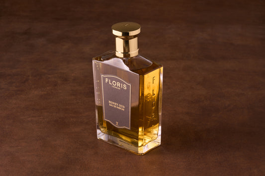 Floris Honey Oud Eau de Parfum in a clear glass bottle with amber liquid and gold cap, displayed on leather, embodies timeless elegance and rich fragrance notes.