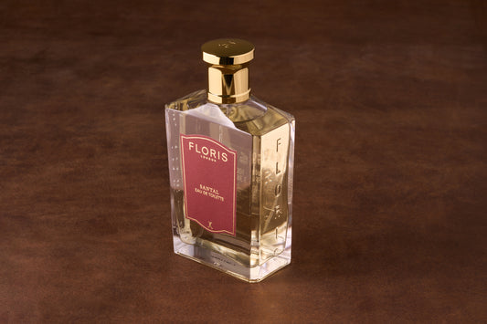 Floris Santal Eau De Toilette in a clear bottle with gold cap and red label, displayed on leather. Features sandalwood, cardamom, nutmeg, frankincense, and musk notes.