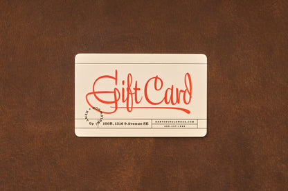 A cream-colored Gift Card from Kent of Inglewood, featuring “Gift Card” prominently in red script, rests on a brown surface. Ideal for shaving gifts or barber services, this card also displays small black text at the bottom with contact details.