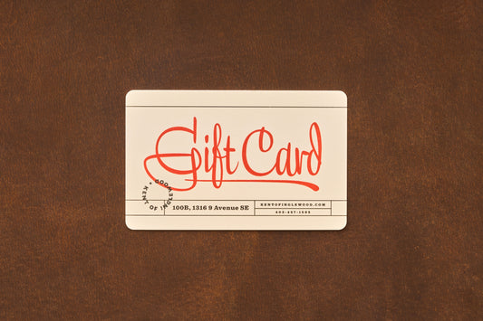 A cream-colored Gift Card from Kent of Inglewood, featuring “Gift Card” prominently in red script, rests on a brown surface. Ideal for shaving gifts or barber services, this card also displays small black text at the bottom with contact details.
