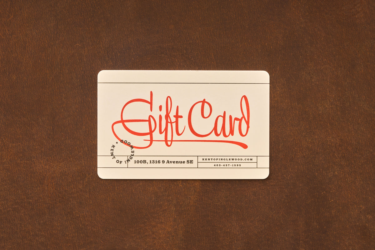 A beige Shaving Class Gift Card, featuring “Gift Card” elegantly scripted in red, rests on a brown surface. Perfect for grooming enthusiasts and those interested in straight razor shaving, this card makes an ideal present for anyone seeking shave class gift cards. The bottom of the card displays an address, website, and phone number in small print.