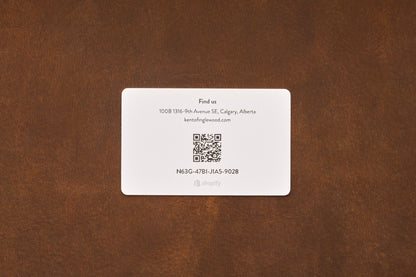 A business card on a brown surface showcases contact details, a QR code, and the code N63G-4T1B-J1A5-9028. Located in Calgary, Alberta, the card also displays an email address and the Shopify logo. It promotes barber services and features the Gift Card from Kent of Inglewood—an ideal choice for anyone searching for distinctive shaving gifts.