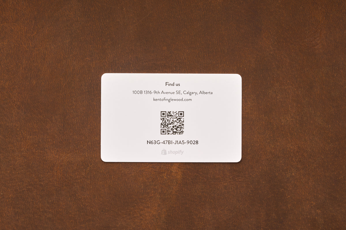 A white business card featuring contact details, a QR code, and the code N63G-4T1B-JIA5-9028 rests on a brown surface. With an address in Calgary, Alberta and an email provided, this card is ideal for fans of straight razor or safety razor shaving. It likely contains information about the Shaving Class Gift Card—excellent for anyone seeking to elevate their grooming experience.